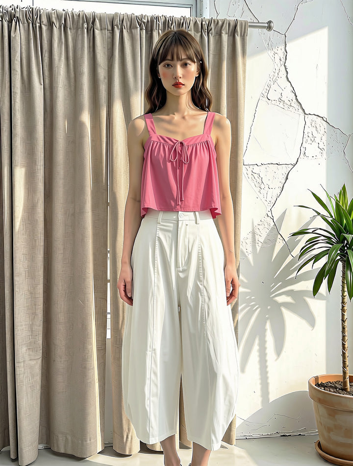A woman stands indoors wearing a pink sleeveless ruffled top and white wide-leg cropped pants, posed in front of beige curtains and a white wall with a potted plant to the right.
