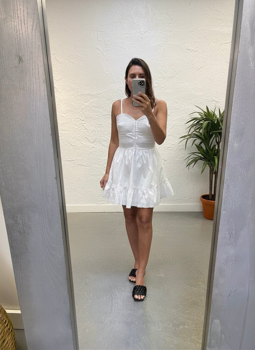 Person wearing a white dress taking a mirror selfie in a room with a plant.