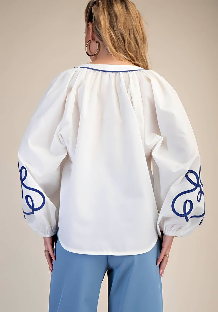 White blouse with blue embroidery on sleeves and neckline worn by a person against a beige background