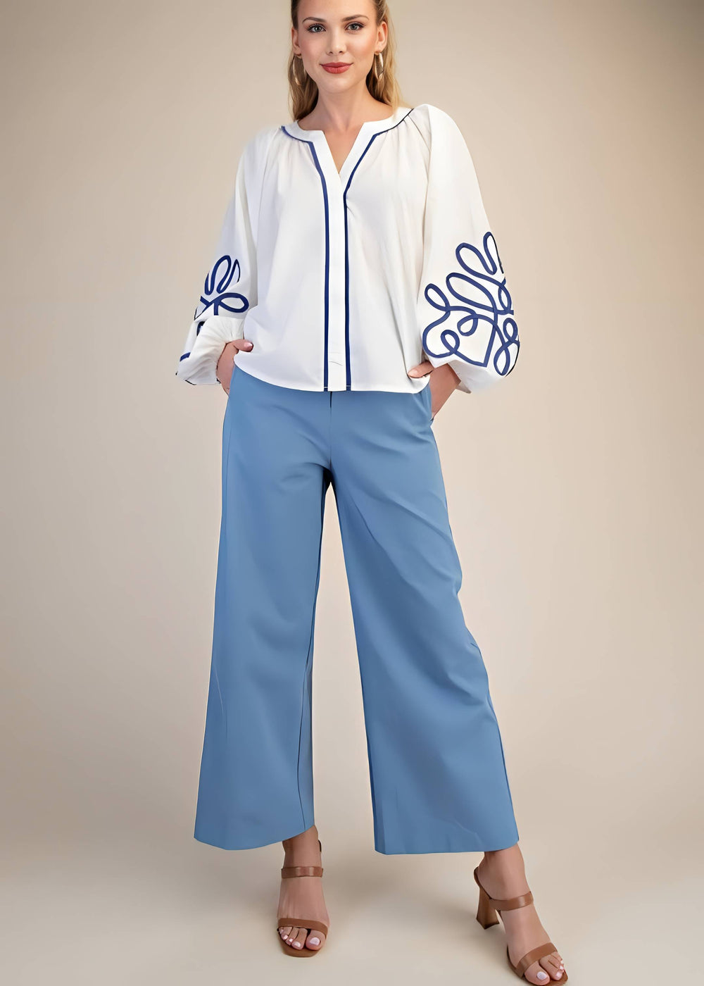 Woman wearing a white blouse with blue embroidery and light blue pants on a beige background