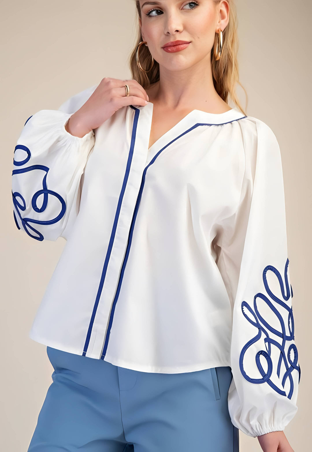 Woman wearing a white blouse with blue embroidery and blue pants against a beige background