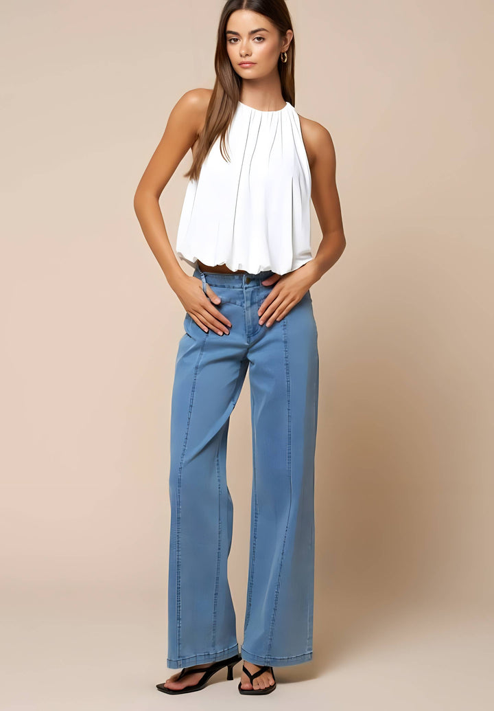 A woman modeling a white sleeveless pleated top and light blue wide-leg jeans against a solid beige studio background.