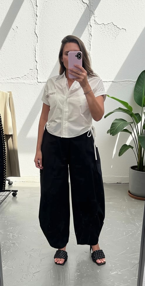A woman takes a mirror selfie wearing a white short-sleeve button-up shirt and black wide-leg pants in a bright room with a white textured wall and a potted plant.