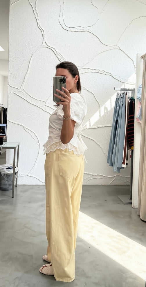 A woman takes a mirror selfie in a clothing boutique, wearing a white ruffled short-sleeve blouse and light yellow wide-leg trousers.