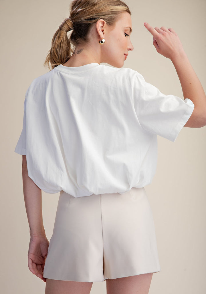A woman shown from the back wearing a loose white short-sleeve top and light beige shorts against a plain beige studio background.