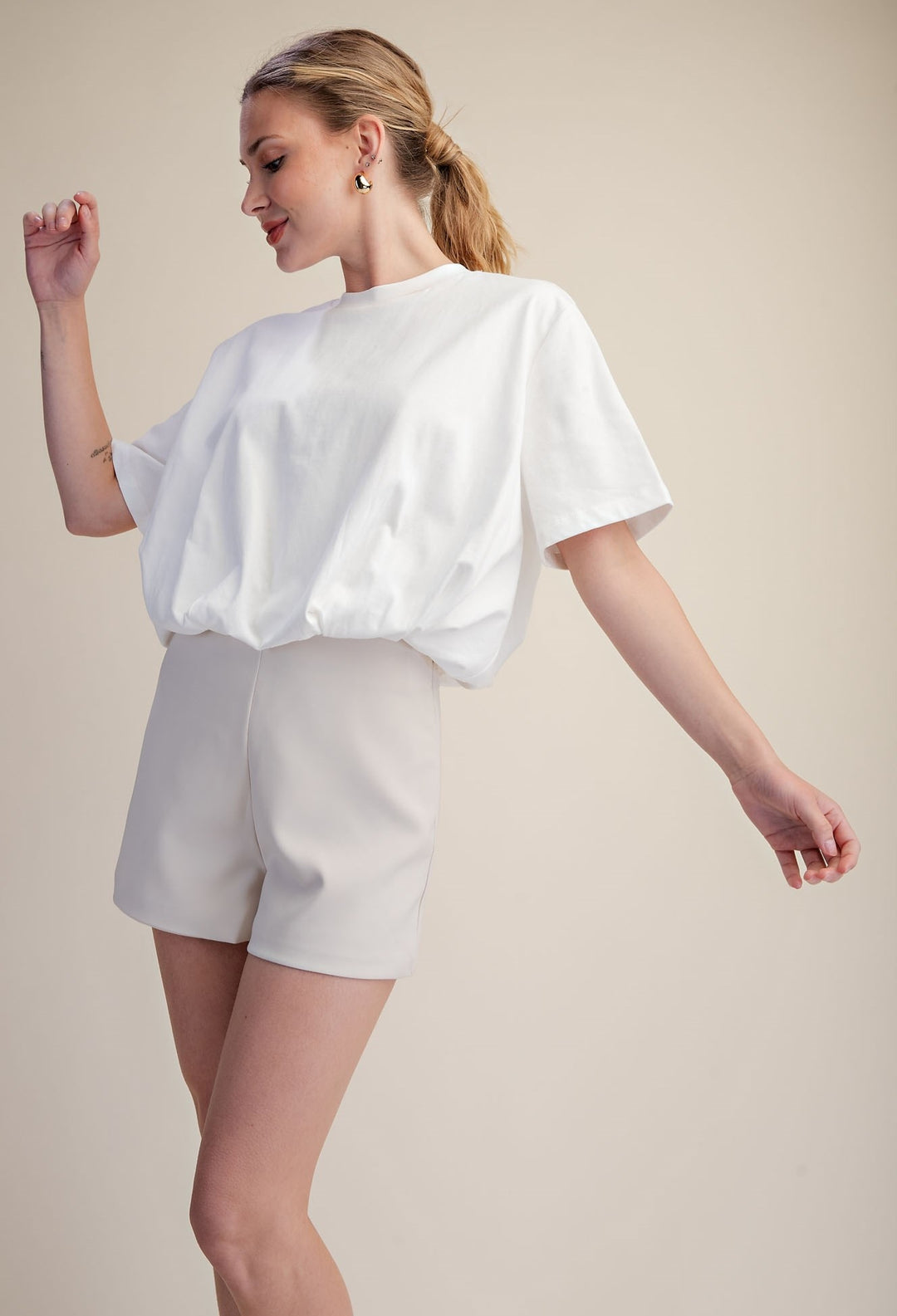 A woman modeling a white short-sleeve top and light beige shorts against a plain beige studio background.