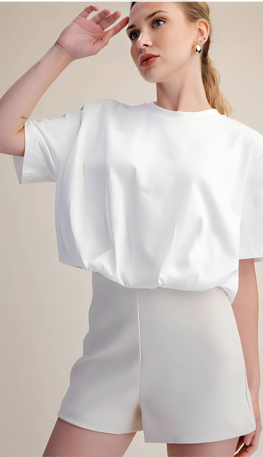 A woman modeling a white short-sleeve top with a voluminous, gathered silhouette and matching white shorts against a plain beige studio background.