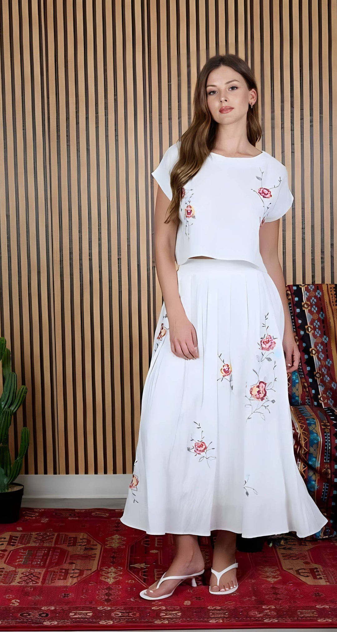 A woman modeling a white two-piece outfit consisting of a short-sleeve crop top and a matching midi skirt, both decorated with pink and red floral embroidery. She is standing in front of a wooden slatted wall and a red patterned rug.