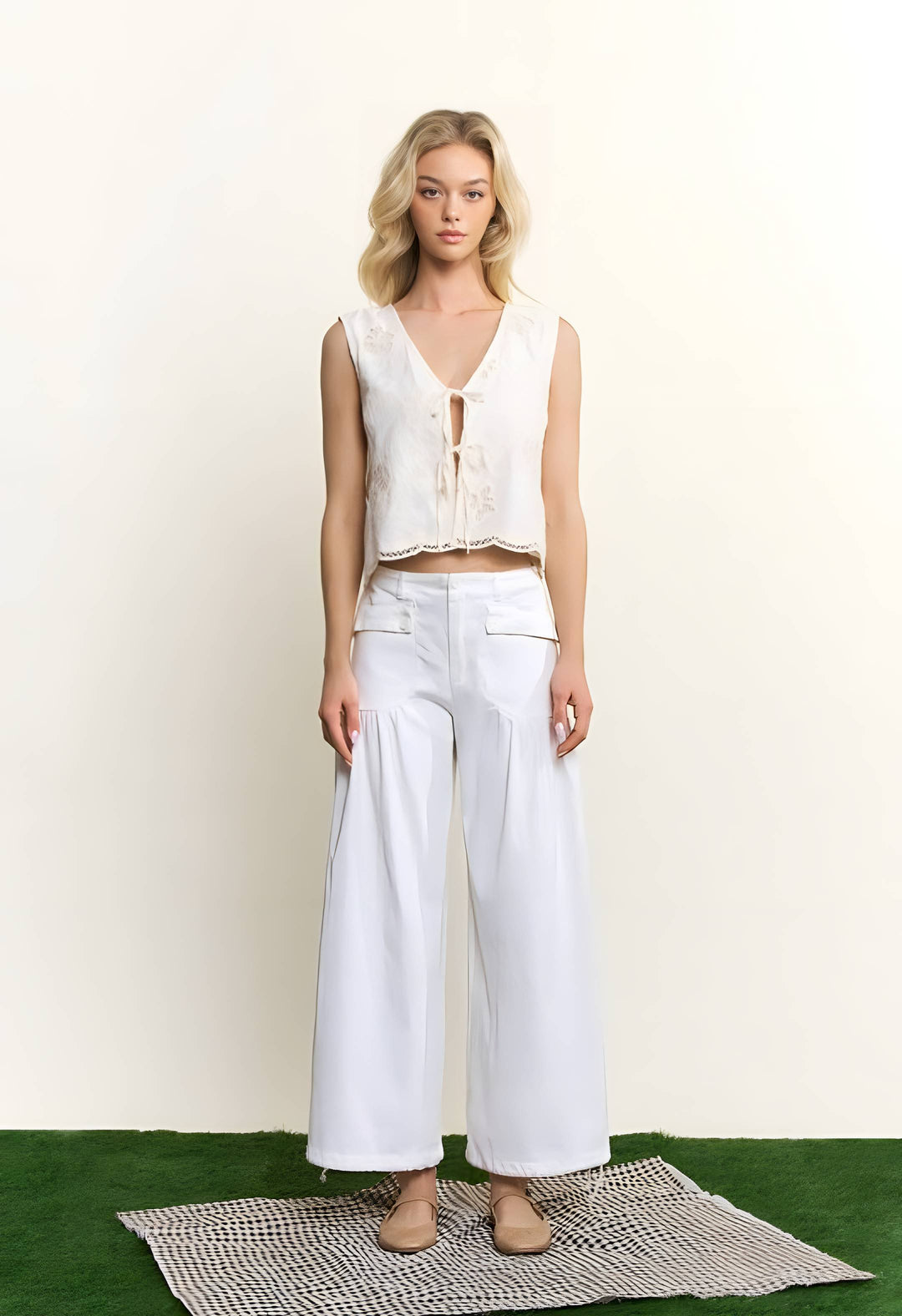A blonde model stands on a textured rug wearing a white sleeveless lace-up crop top and matching white wide-leg pants, set against a plain off-white wall with green turf at the bottom.