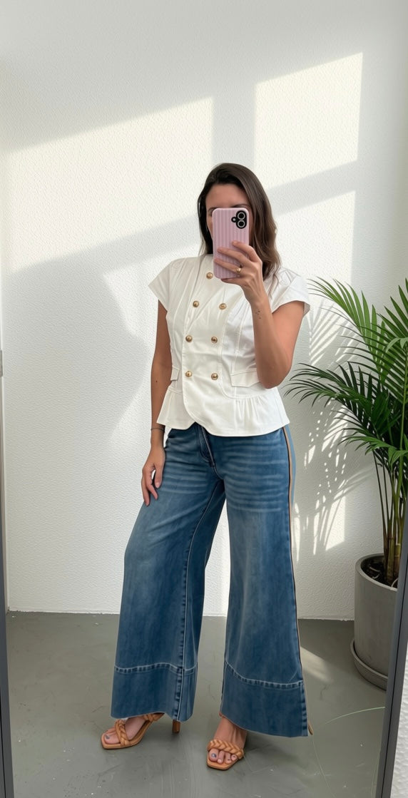 A woman takes a mirror selfie wearing a white double-breasted short-sleeve blouse with gold buttons and wide-leg blue denim jeans, paired with beige heeled sandals, in a bright room with a potted plant.