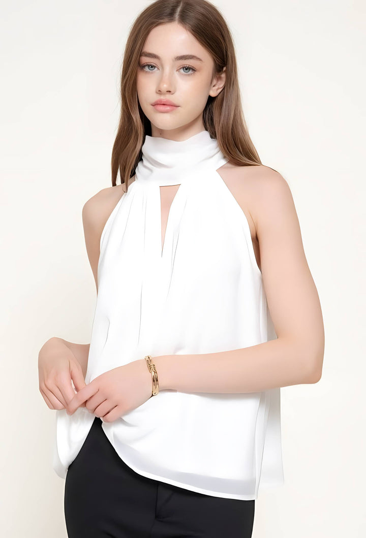 A woman modeling a white sleeveless high-neck blouse with a keyhole cutout and draped tie detail, paired with black pants against a plain light background.