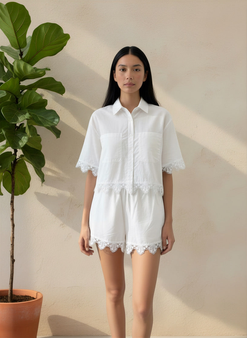 Woman wearing a white outfit with lace details standing next to a potted plant against a beige wall.