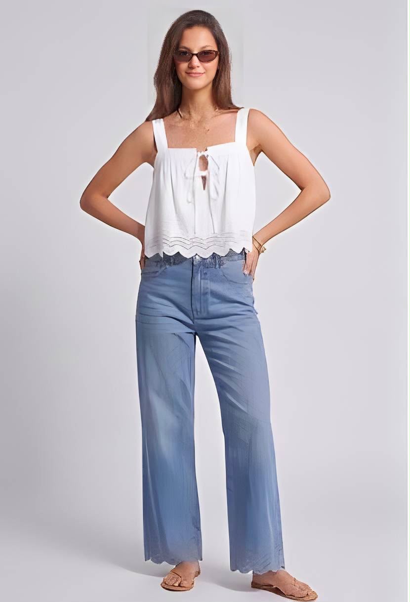 A woman modeling a white sleeveless top with a scalloped hem and tie detail, paired with wide-leg light blue jeans and tan sandals, against a light gray studio background.