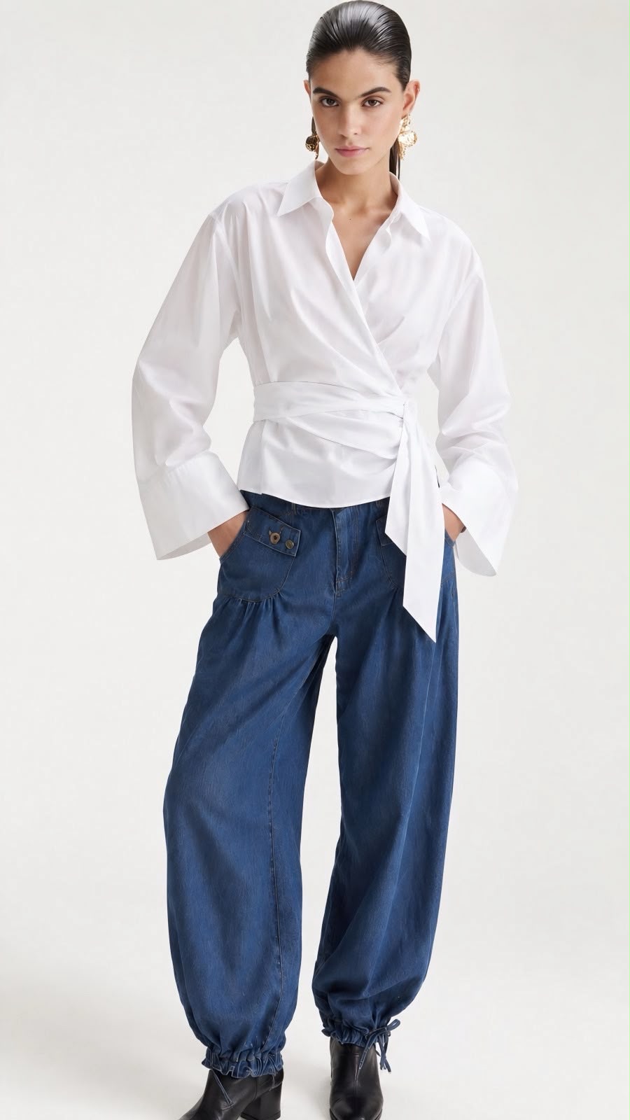 A model wearing a white long-sleeve wrap-style shirt tied at the waist and dark blue wide-leg pants, standing against a plain light background.