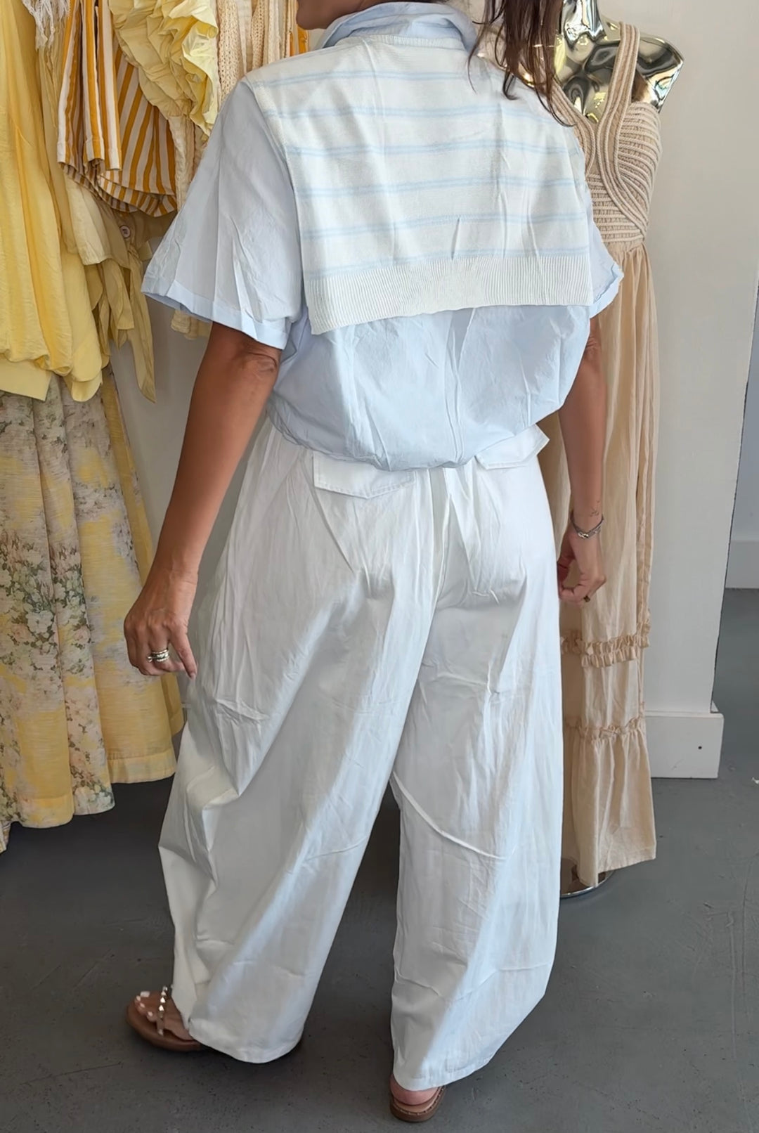 A person shown from the back wearing a white short-sleeve collared shirt with a wide collar and matching wide-leg pants, standing in a boutique with other dresses in the background.