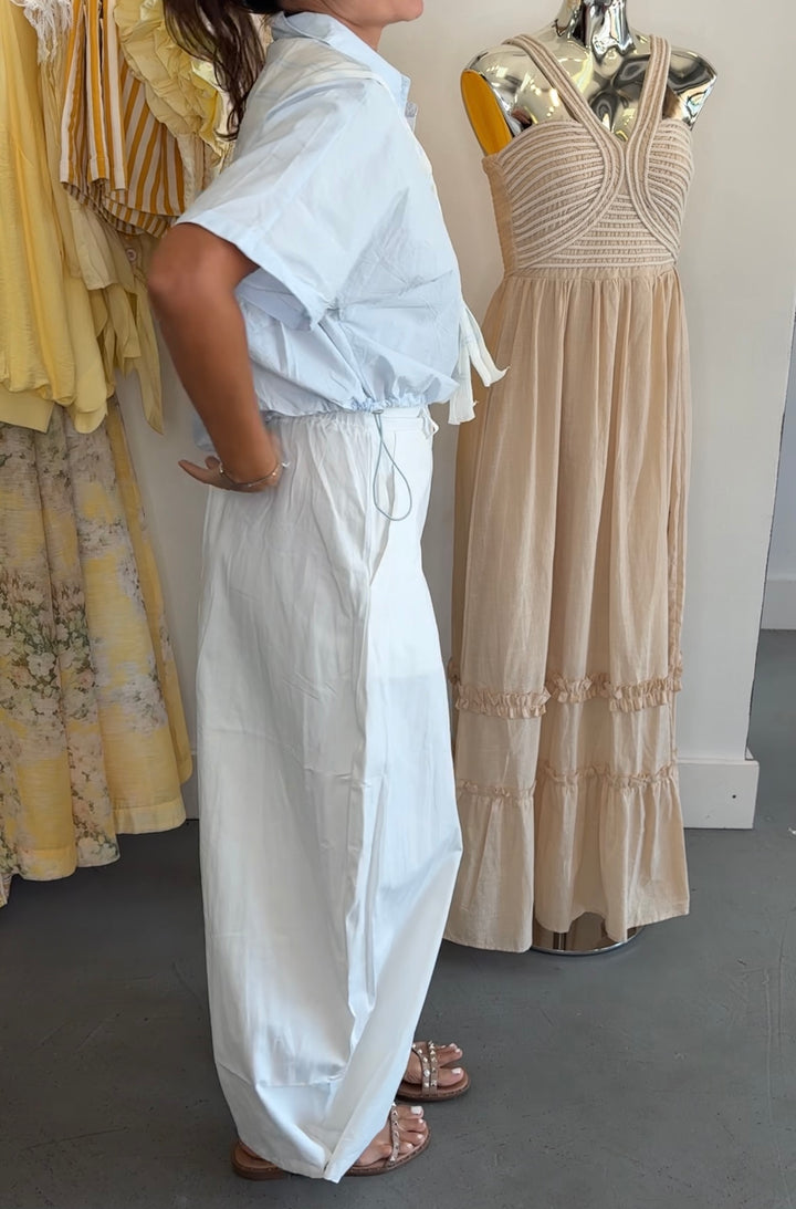 A model wearing a light blue short-sleeve button-up shirt and white wide-leg trousers stands in a clothing store next to a beige maxi dress displayed on a mannequin.