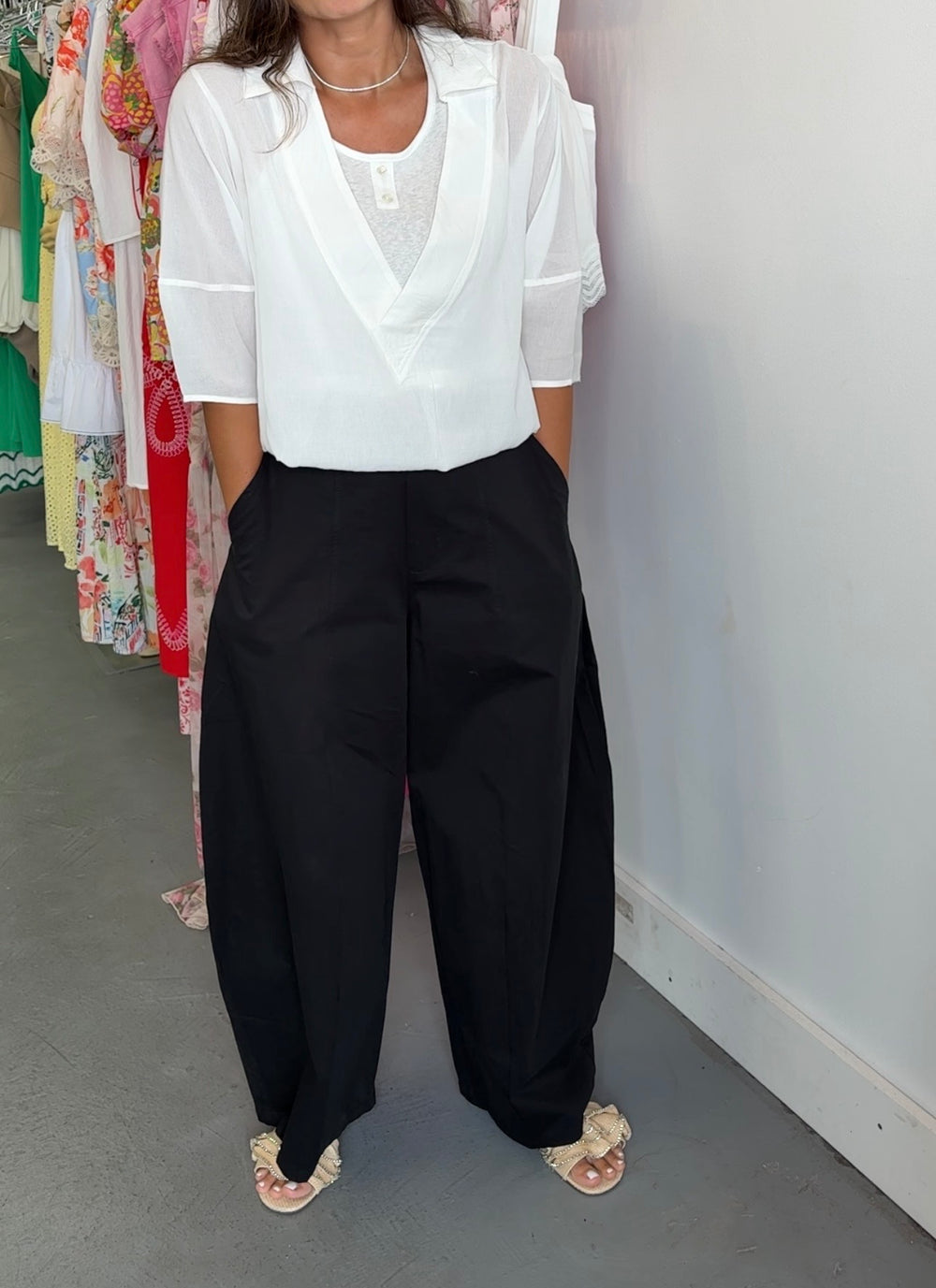A person wearing a white short-sleeve blouse with a V-neck and black wide-leg pants, standing indoors against a white wall with clothing racks visible in the background.