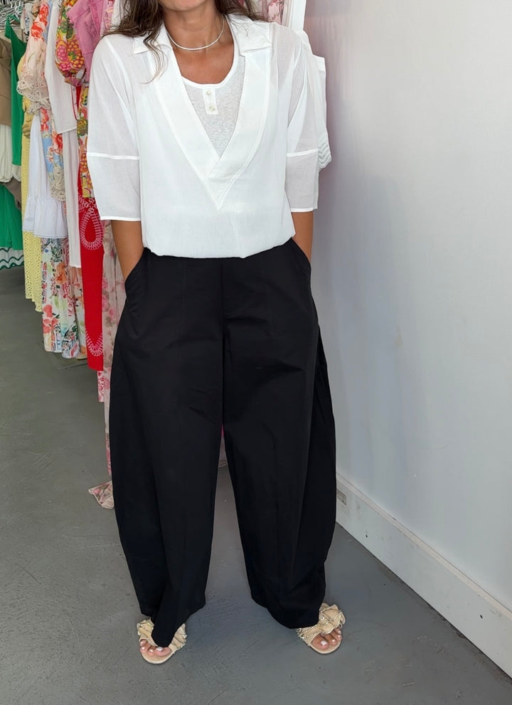A person wearing a white short-sleeve blouse with a V-neck and black wide-leg pants, standing indoors against a white wall with clothing racks visible in the background.