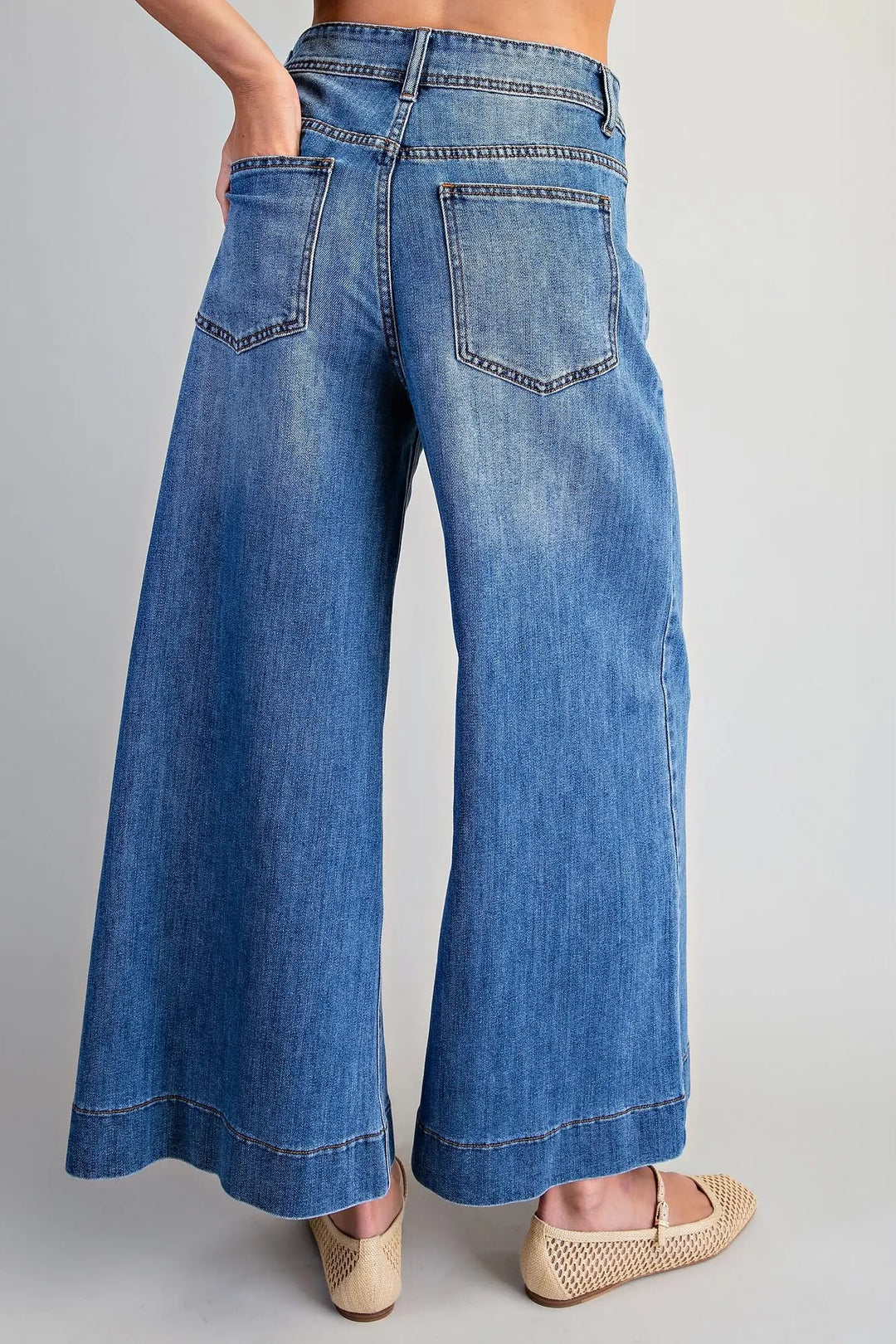 A person wearing blue wide-leg denim jeans with a faded wash, shown from the back against a light gray studio background. The jeans have a high waist, back pockets, and a cropped leg length.