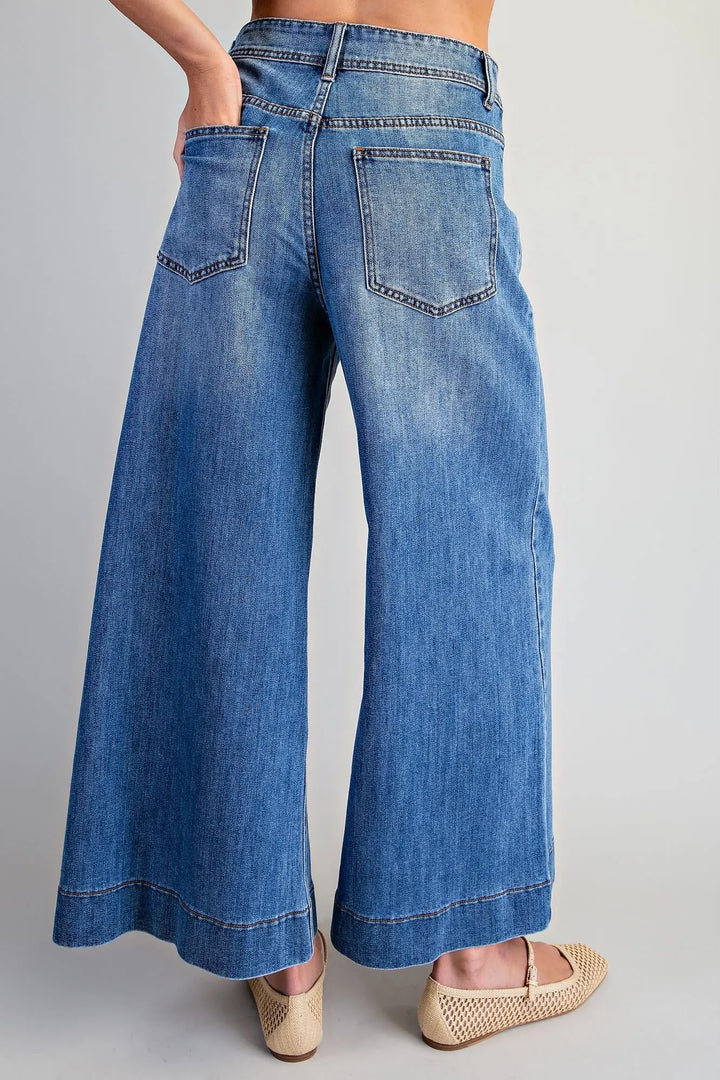 A person wearing blue wide-leg denim jeans with a faded wash, shown from the back against a light gray studio background. The jeans have a high waist, back pockets, and a cropped leg length.