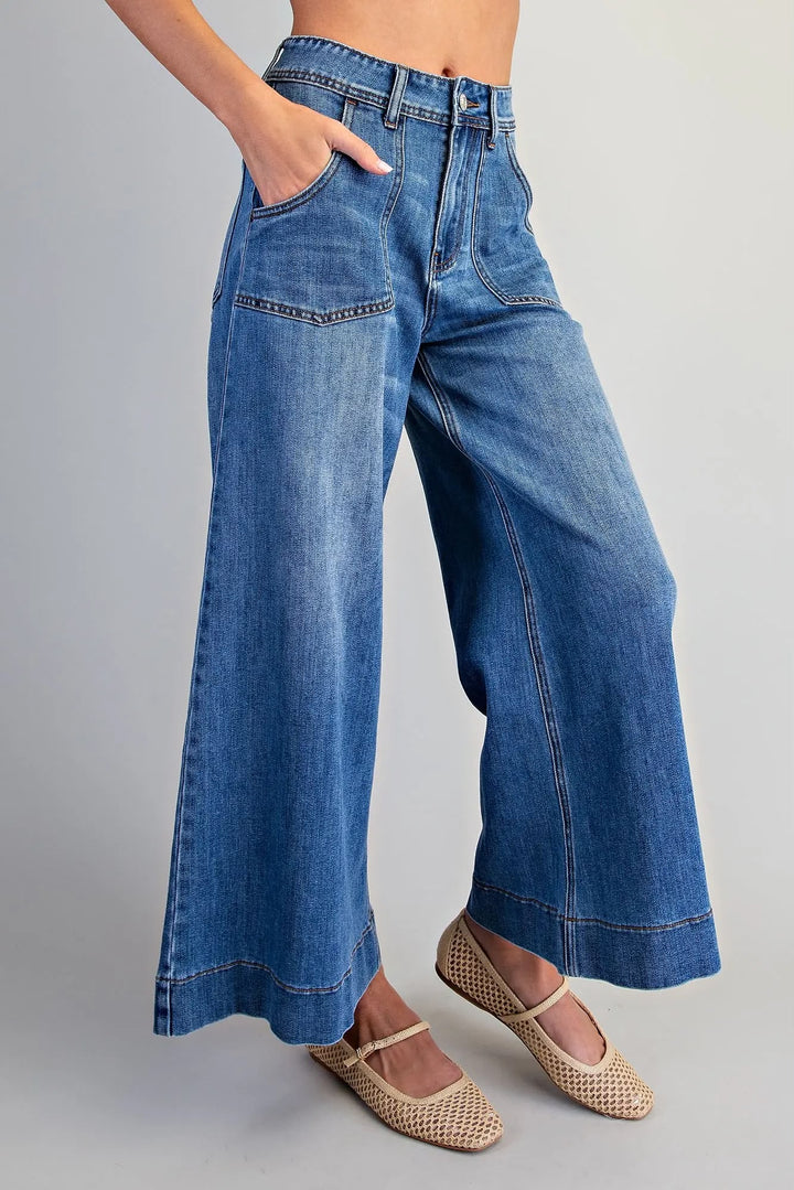 A model wearing high-waisted, wide-leg blue denim jeans with a relaxed fit and cuffed hems, paired with beige woven flat shoes against a neutral gray background.