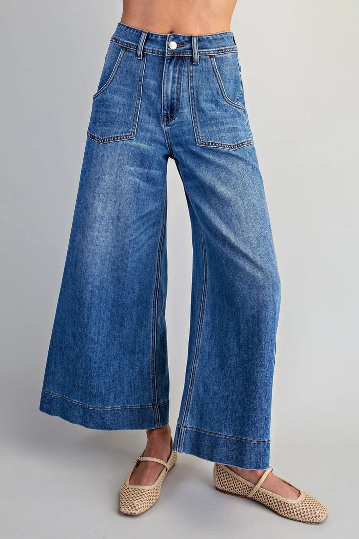 A person wearing blue wide-leg denim jeans with a faded wash and tan woven flat shoes, shown from the waist down against a light gray studio background.