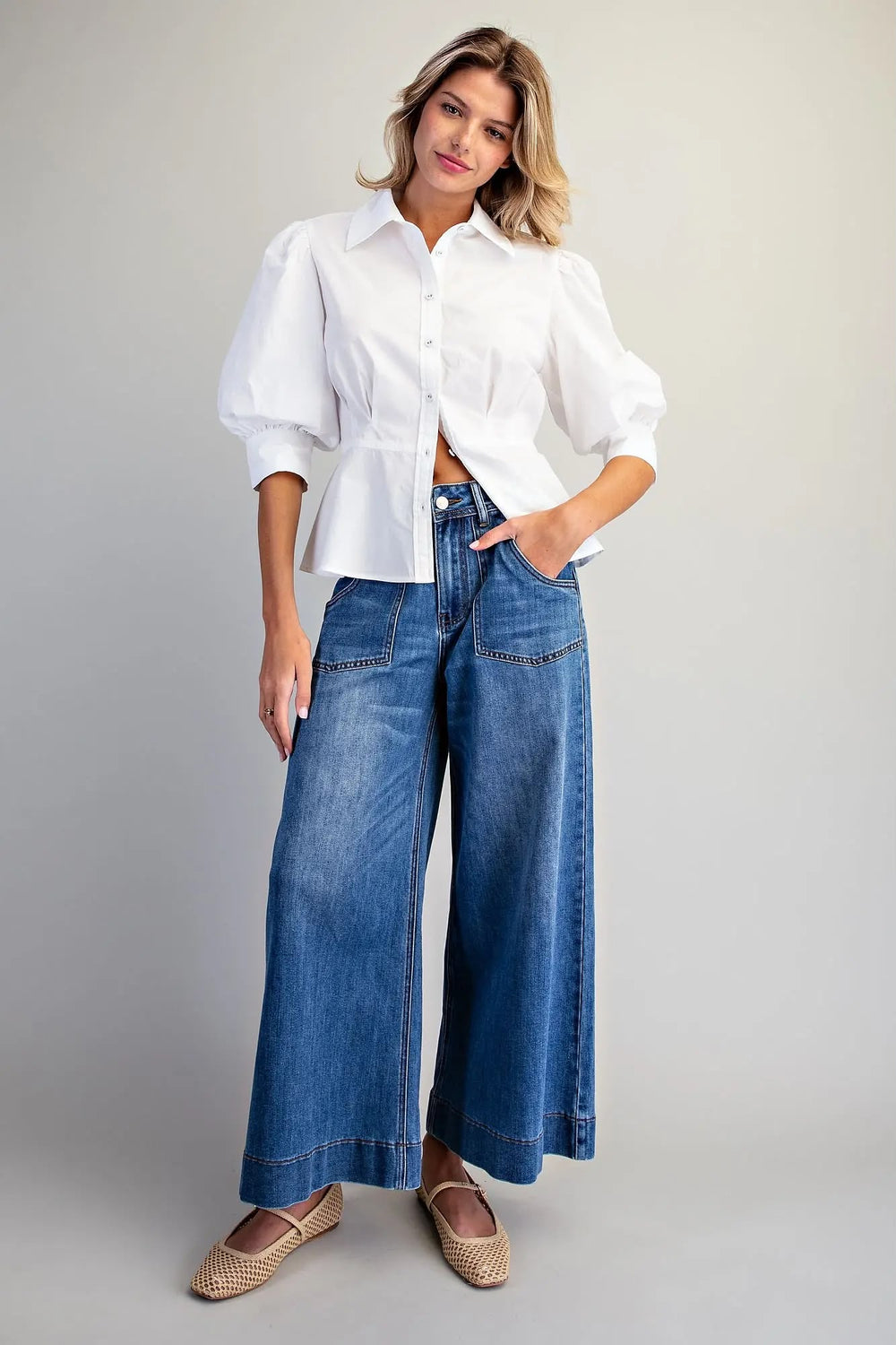 A woman modeling a white long-sleeve blouse with voluminous puff sleeves and blue wide-leg denim jeans, paired with woven espadrille flats, against a neutral light gray studio background.