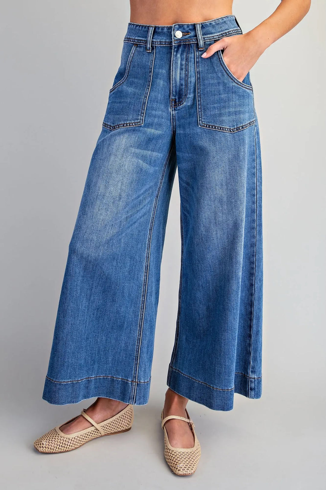 A model wearing high-waisted, wide-leg blue denim jeans with front patch pockets and rolled hems, paired with woven flat shoes, against a neutral gray studio background.