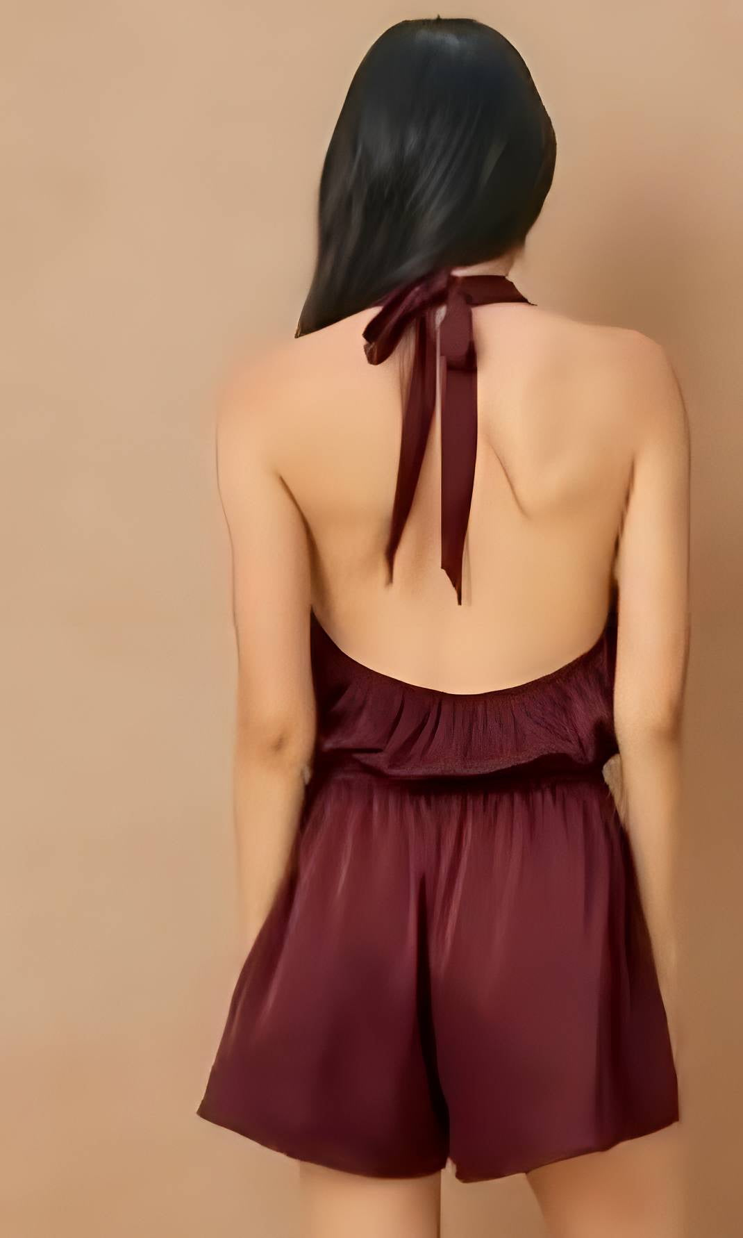 A woman shown from the back wearing a sleeveless burgundy halter romper with a tied back and ruffled waist, against a plain beige studio background.
