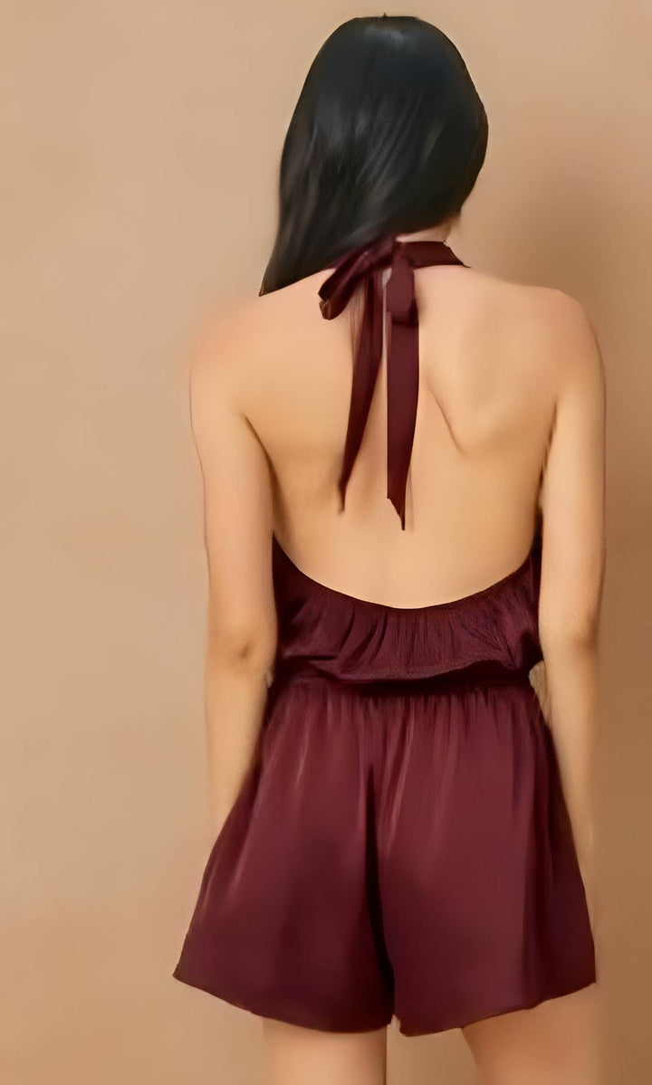 A woman shown from the back wearing a sleeveless burgundy halter romper with a tied back and ruffled waist, against a plain beige studio background.