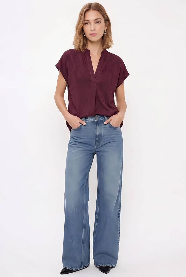A woman modeling a short-sleeve burgundy blouse and wide-leg blue jeans against a plain white studio background.