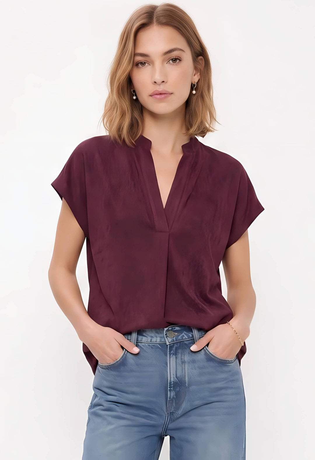 A woman wearing a short-sleeve, deep burgundy blouse with a split V-neckline, paired with light blue jeans, against a plain white studio background.