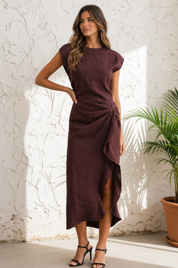 A woman modeling a sleeveless, high-neck, dark plum midi dress with a draped side detail and a front slit, standing against a textured white wall next to a potted plant.