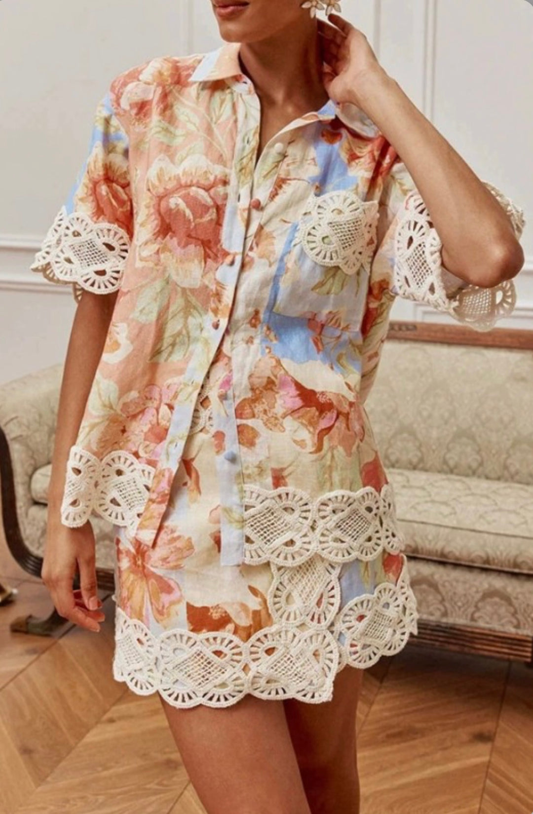 A woman wearing a short-sleeve button-up shirt dress with a pastel floral print and white crochet lace trim, standing in an indoor room with a sofa and wood floor.