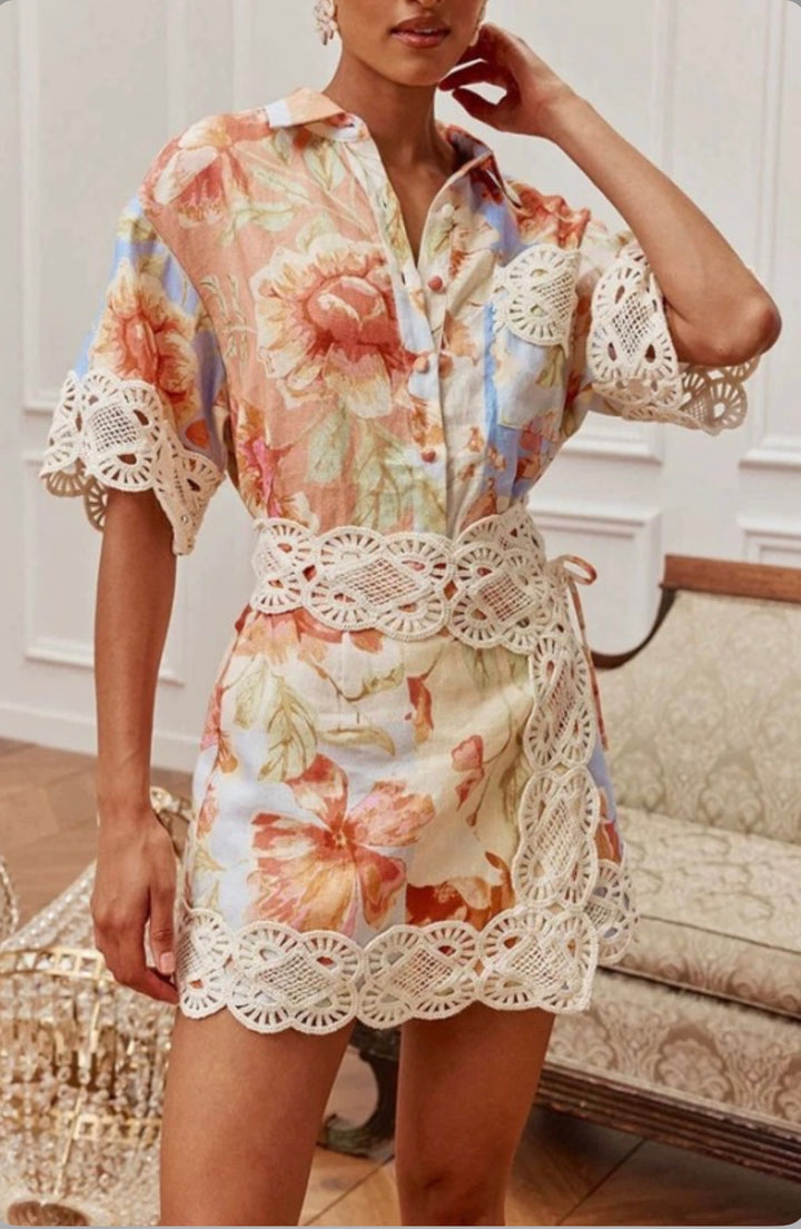 A woman models a short floral print shirt dress with cream lace trim and a lace tie belt, standing in an indoor room with a patterned sofa in the background.
