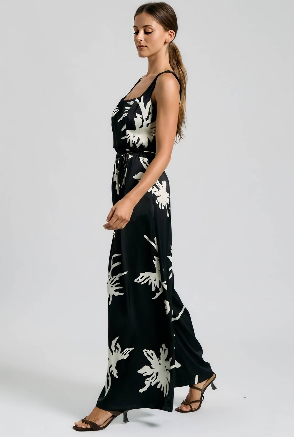 A woman modeling a black sleeveless maxi dress with a white floral print, shown in profile against a light gray studio background.