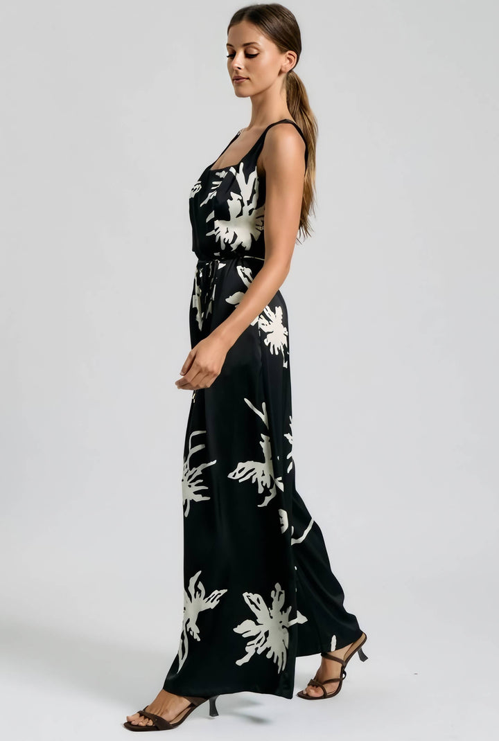 A woman modeling a black sleeveless maxi dress with a white floral print, shown in profile against a light gray studio background.